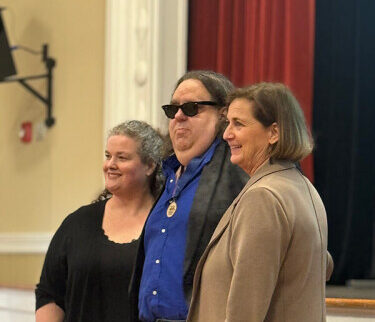 Standing left-to-right are Jennifer Stuckey, Joey Stuckey, and Julie Wilkerson (director of Macon Arts Alliance). Joey is wearing sunglasses, a blue shirt, a black jacket, and, most importantly, the medal he was presented for winning the Macon Arts Alliance Cultural Award. The event took place at the Mill Hill Arts Center in Macon, Georgia.